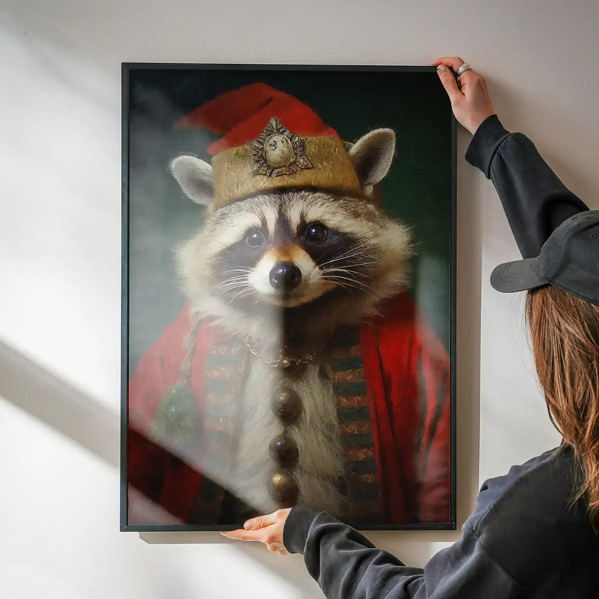 Framed Christmas raccoon portrait print in a red costume and gold hat, held against a white wall.